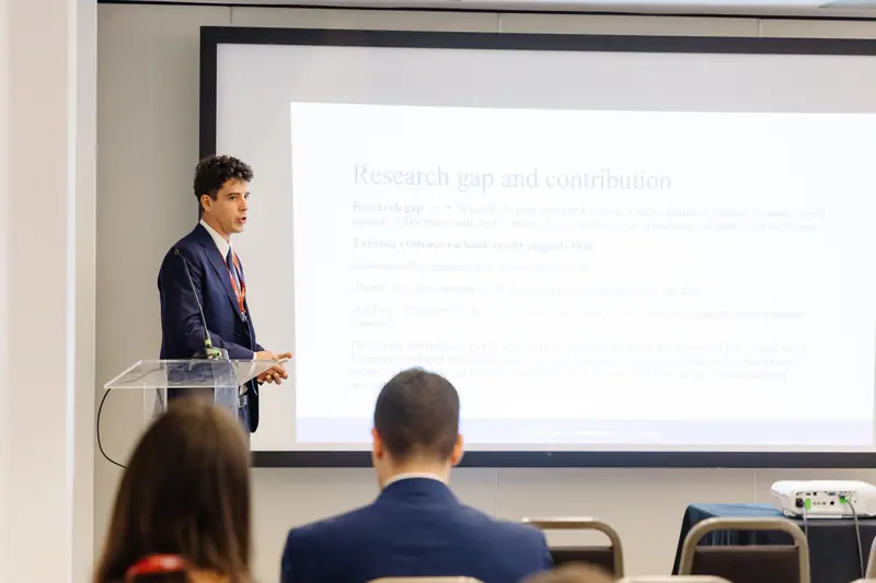 An author presenting a paper beside a slide titled Research gap and contribution, at the 2026 edition.