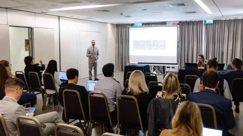 A parallel session at the 2026 edition: a presenter beside the screen and participants seated in a meeting room.